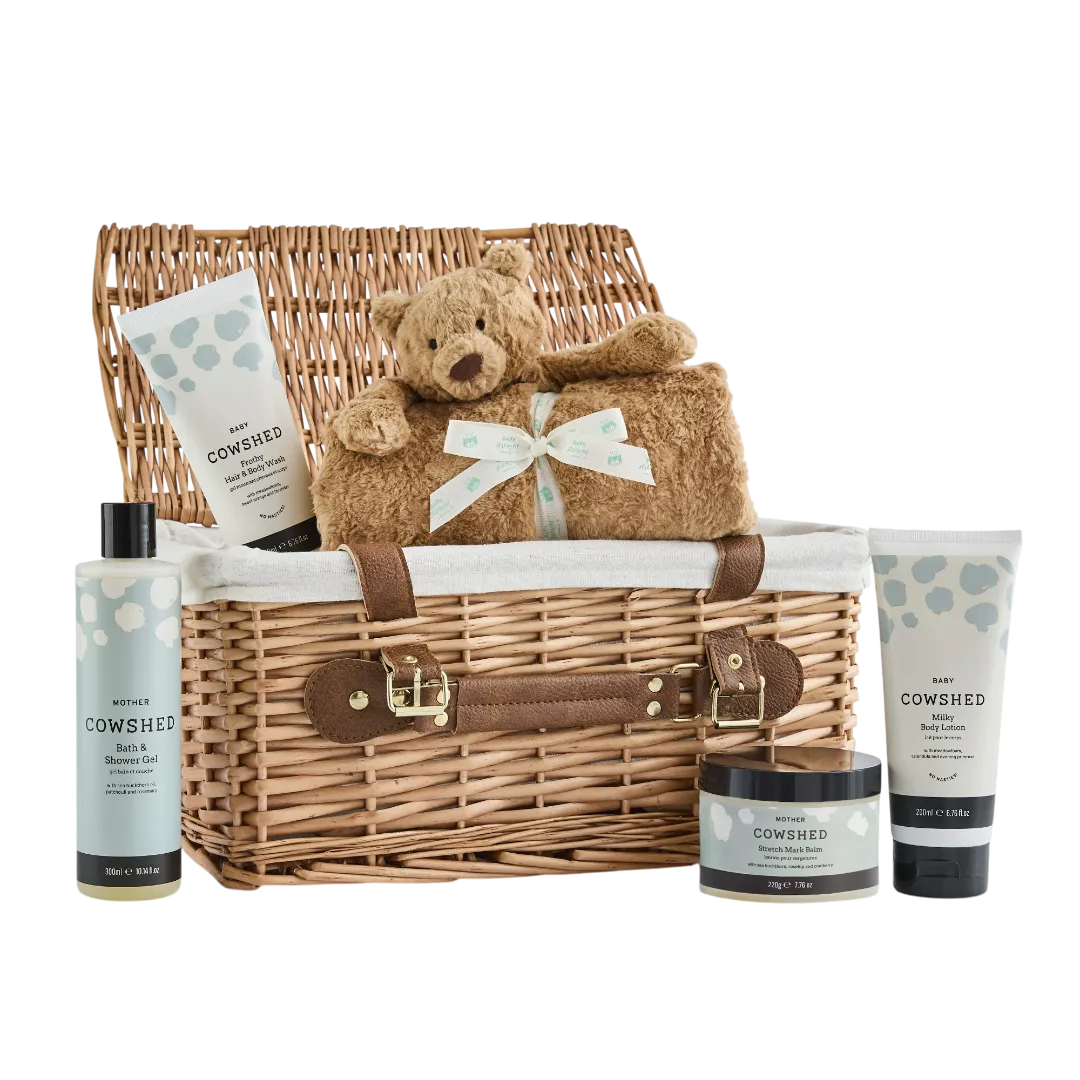 Cowshed baby gift set with wicker basket, teddy bear, and baby products on a white background