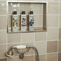 Three bottles of Cowshed products on a tiled wall shelf above a shower head.