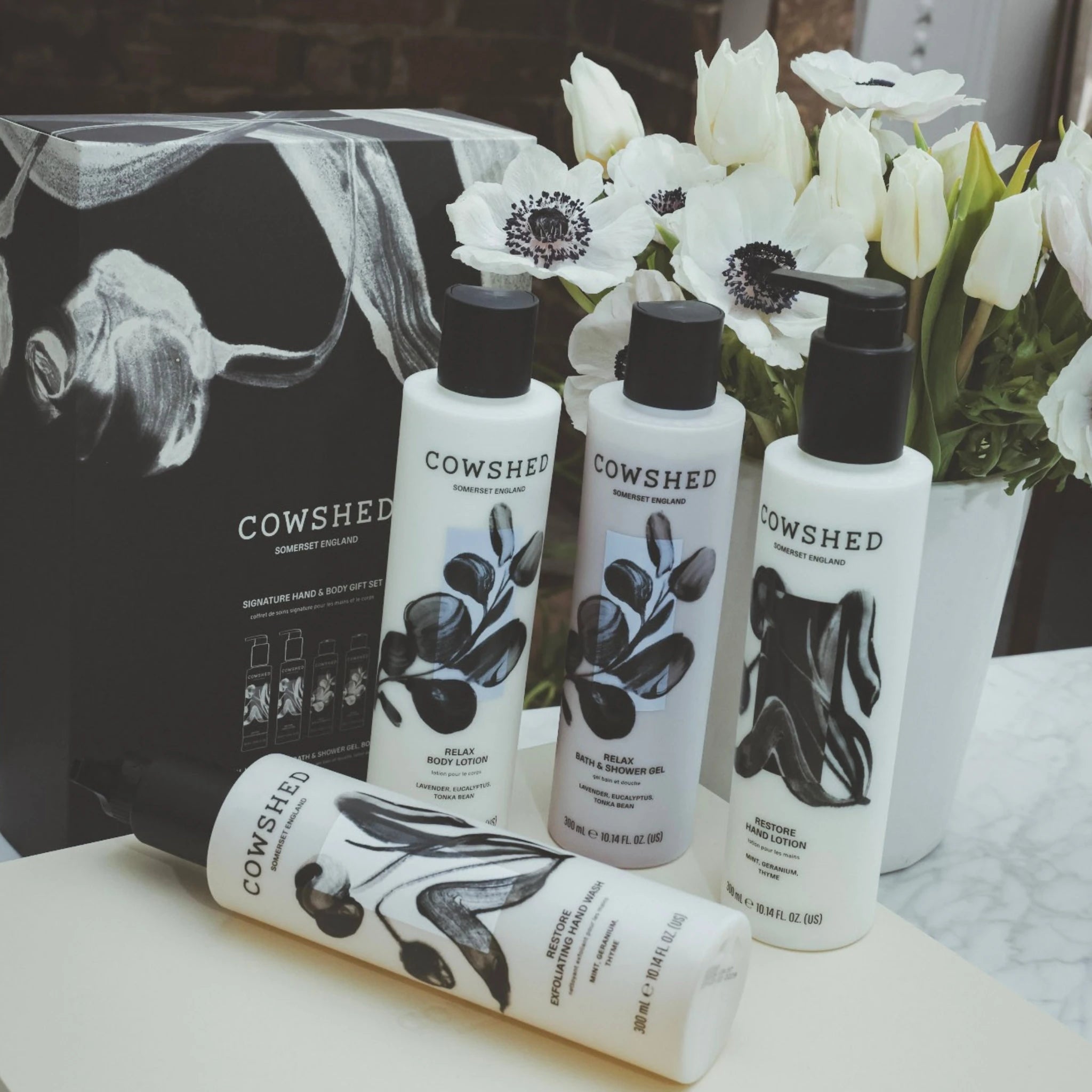 Cowshed skincare products on a table with a decorative box and flowers in the background