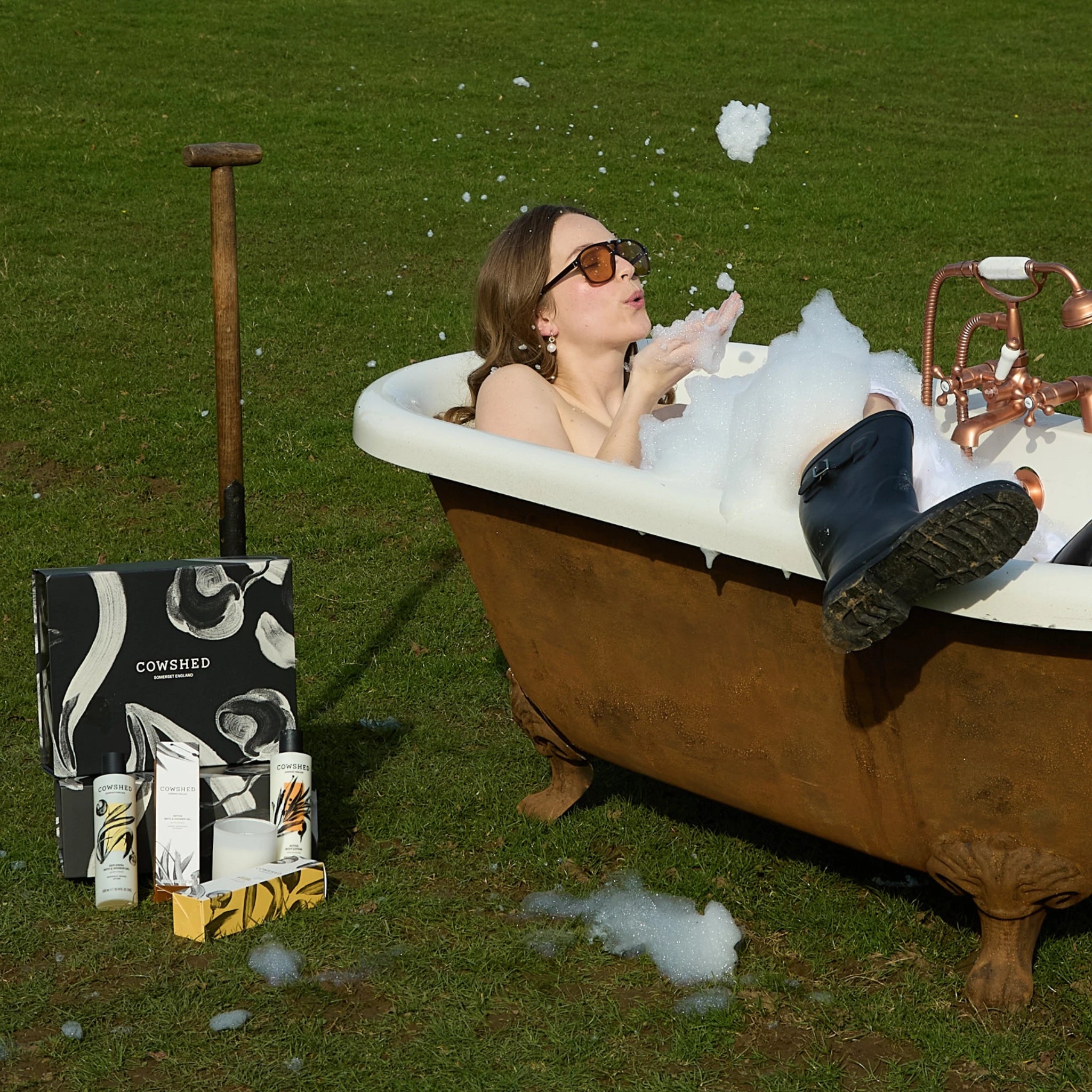 Person taking a bubble bath outdoors with a Cowshed product display nearby.