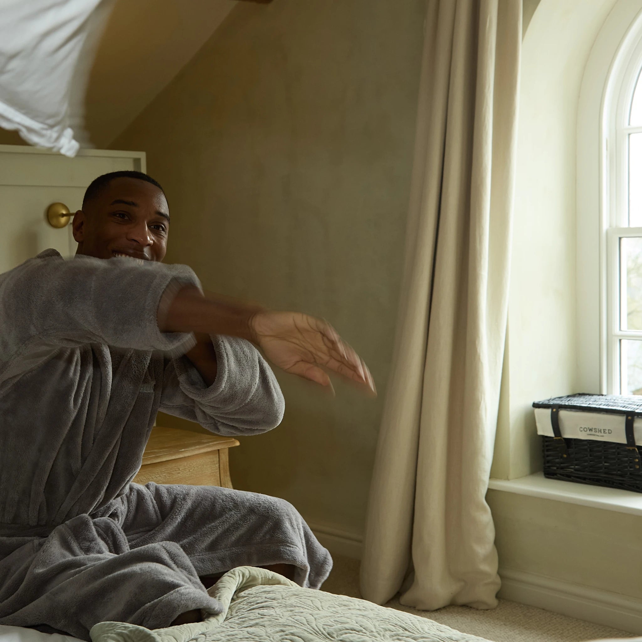 Man in a gray robe sitting on a bed in a bedroom with a window and curtains.