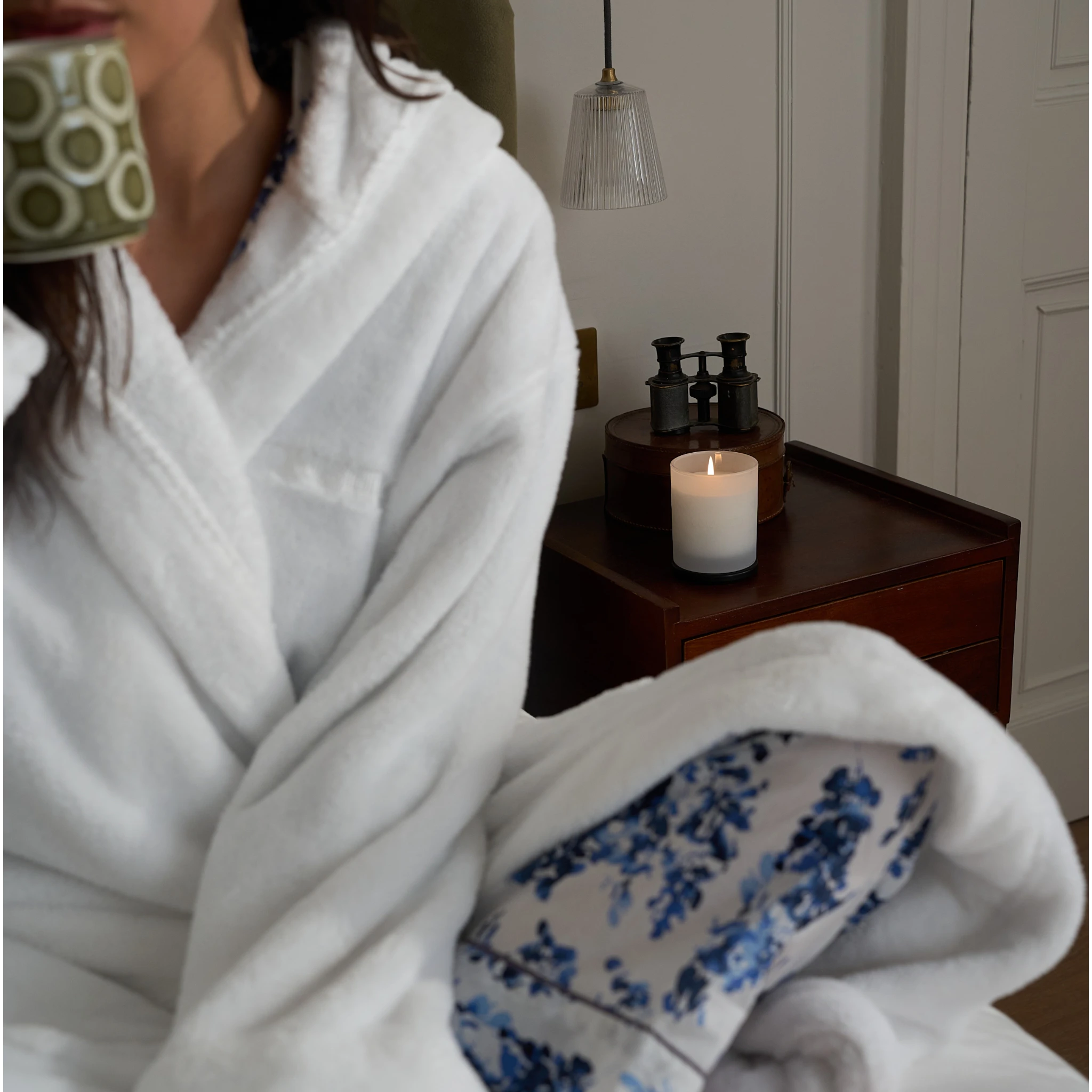 Person in a white robe holding a mug, sitting by a candlelit nightstand.