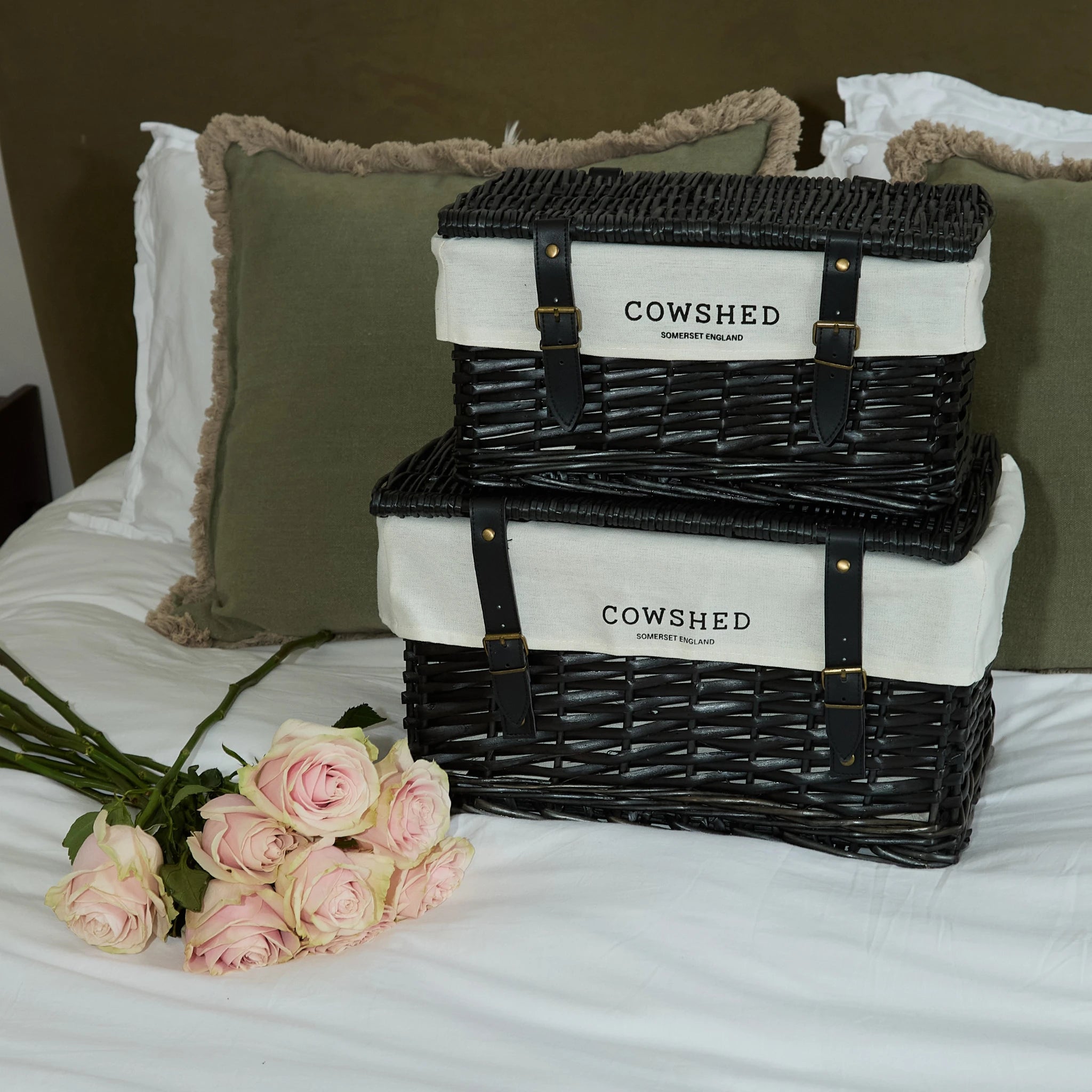 Stack of wicker baskets with 'Cowshed' branding on a bed with pink roses.