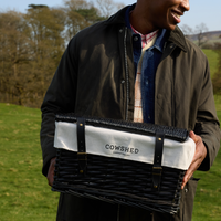 Person holding a black and white hamper with 'Cowshed' branding outdoors.