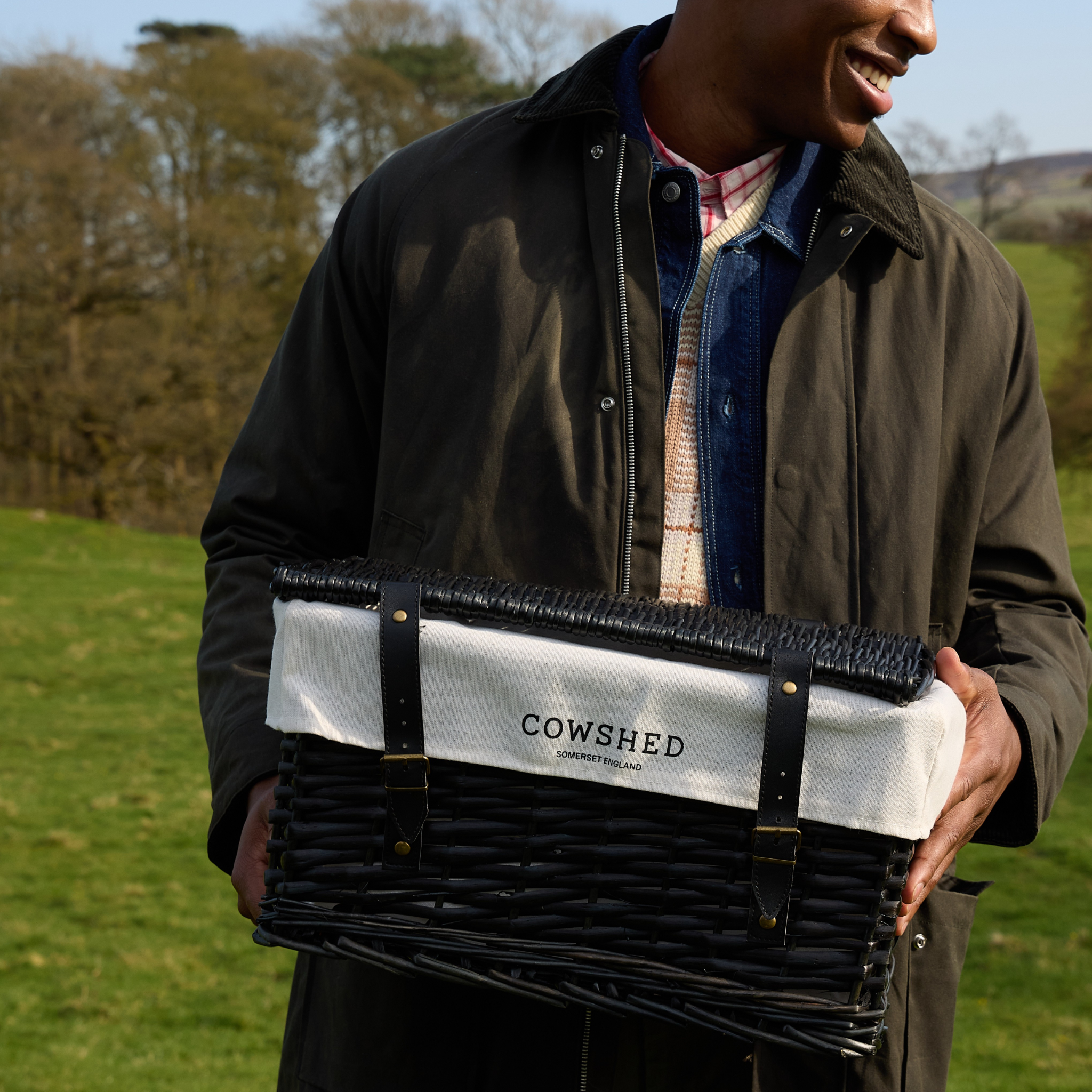 Person holding a black and white hamper with 'Cowshed' branding outdoors.