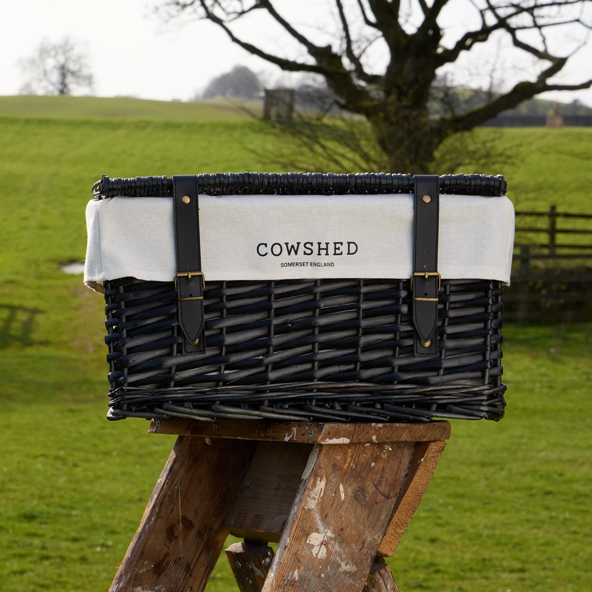 Black wicker basket with 'Cowshed' branding on a wooden stand in a grassy field.