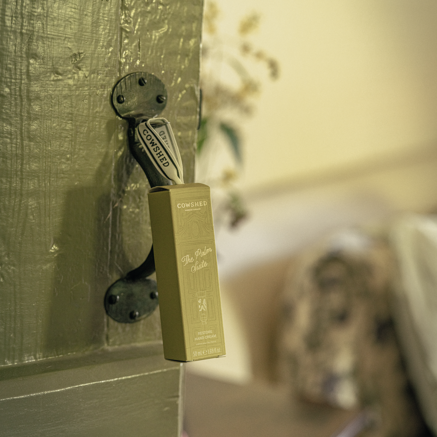 A Cowshed hand cream bauble attached to a rustic green door, with its matching olive-green product box hanging from it in a softly blurred indoor setting.