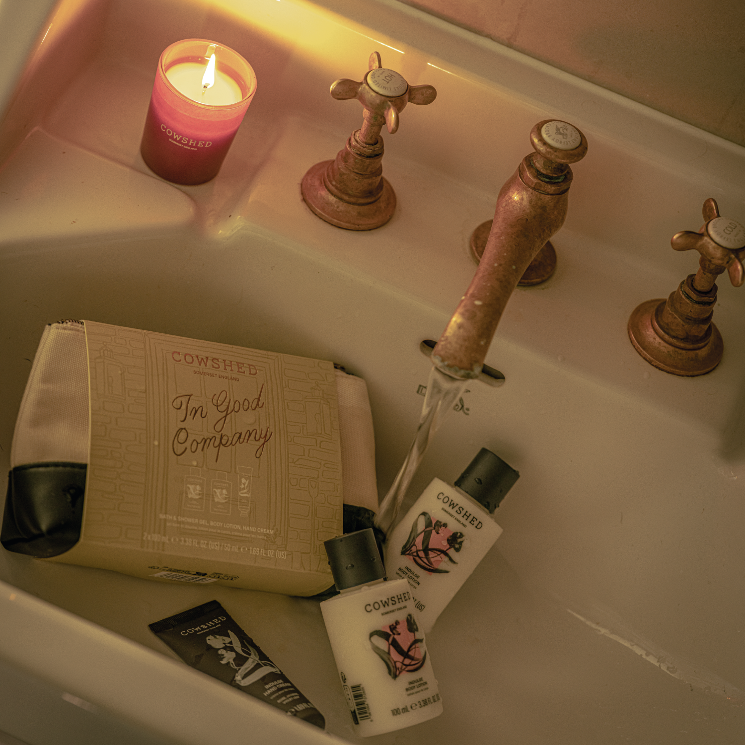 A warm, atmospheric bathroom scene showing a sink with running water, a lit pink candle, and several Cowshed products including a shower gel, body lotion, and a tube of hand cream, all arranged around the faucet.
