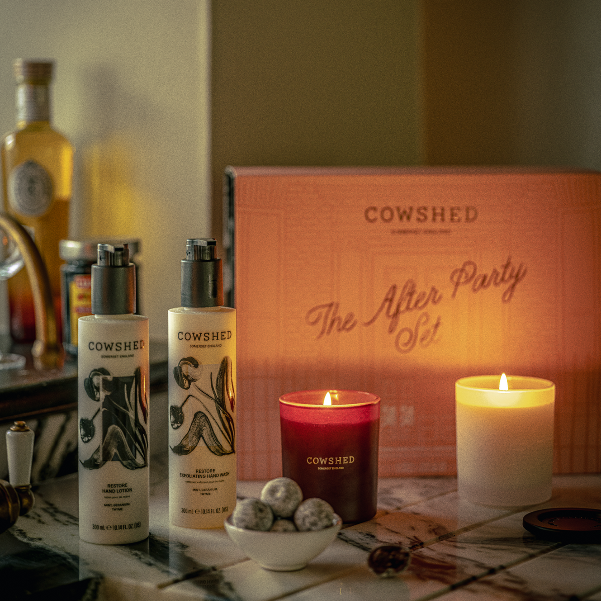 Cowshed 'The After Party Set' gift box displayed on a marble countertop with matching products including candles, hand wash and lotion, alongside drinks and snacks in a warm, dimly lit bar-like setting.