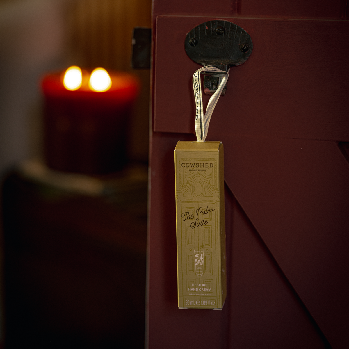 A Cowshed hand cream bauble Christmas gift hangs from a black metal latch on a red barn door, with a warm, softly blurred interior background.