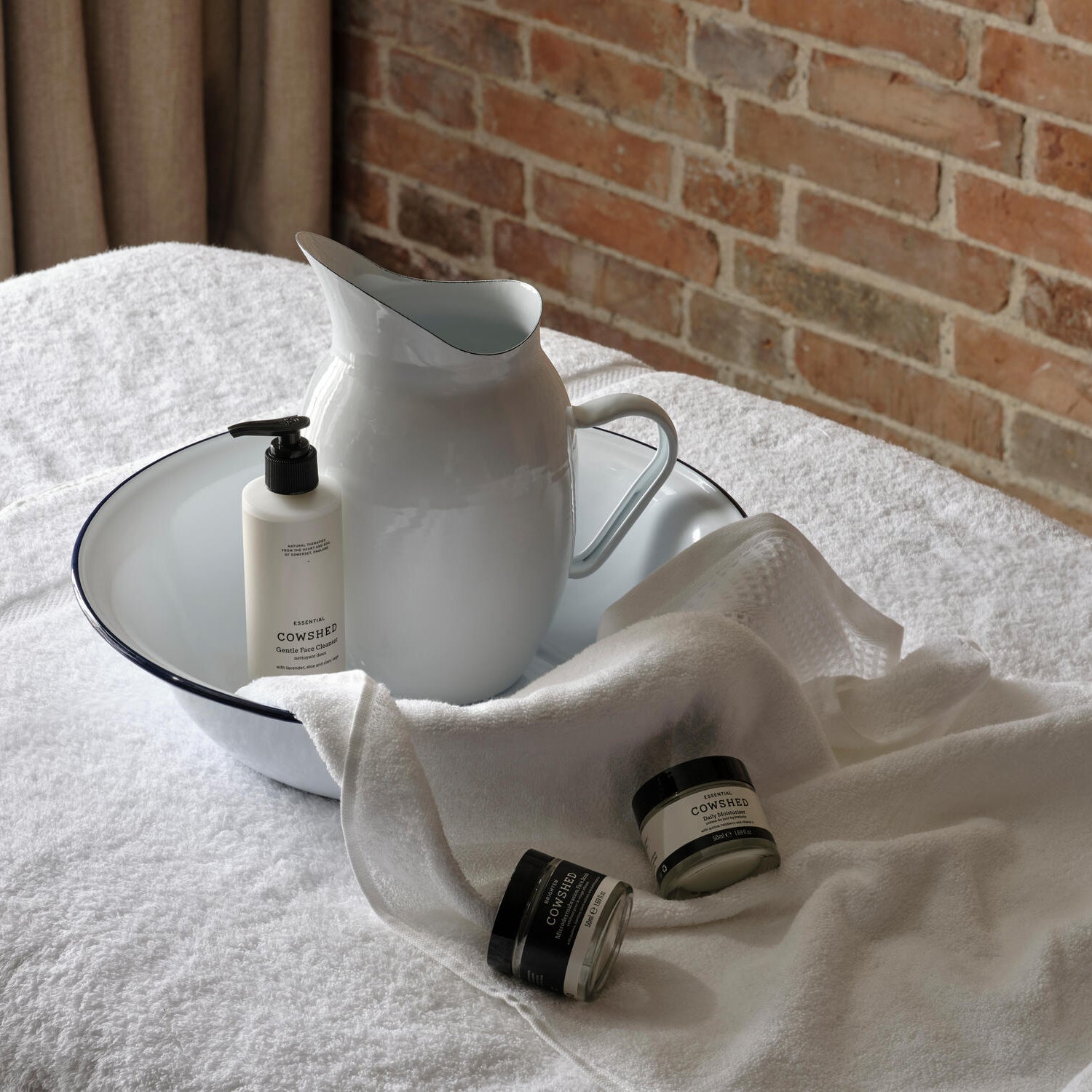 White pitcher and bottles on a white towel with a brick wall background