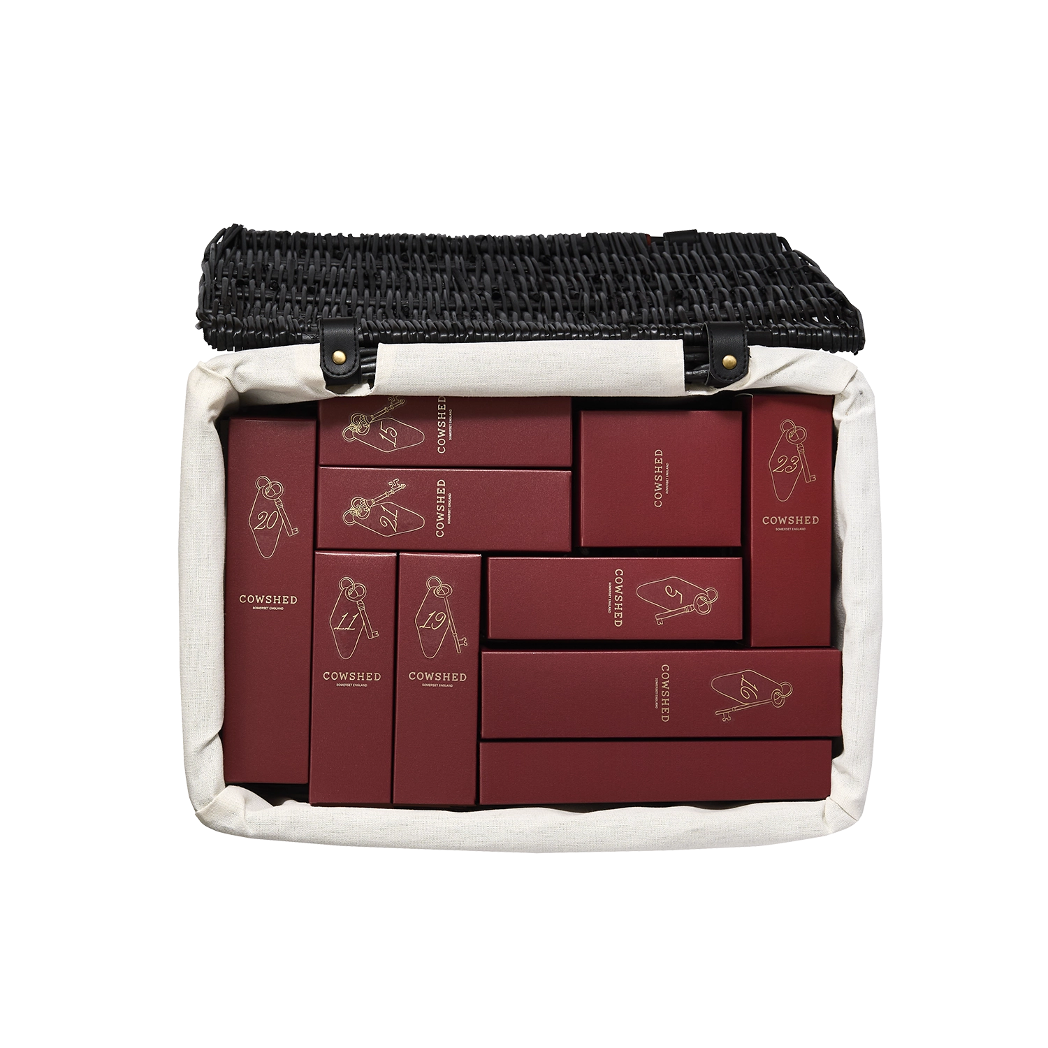 A wicker black hamper basket with a white fabric lining, shown open and filled with multiple burgundy boxes branded Cowshed. A small vintage-style key rests on top of the boxes.