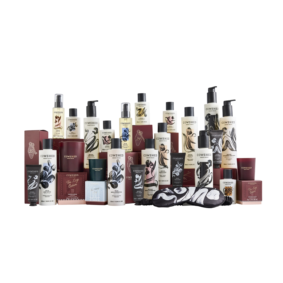 A collection of Cowshed haircare, body care, home fragrance and beauty products arranged together on a plain white background, including bottles, jars, boxes, and a black-and-white patterned eye mask.