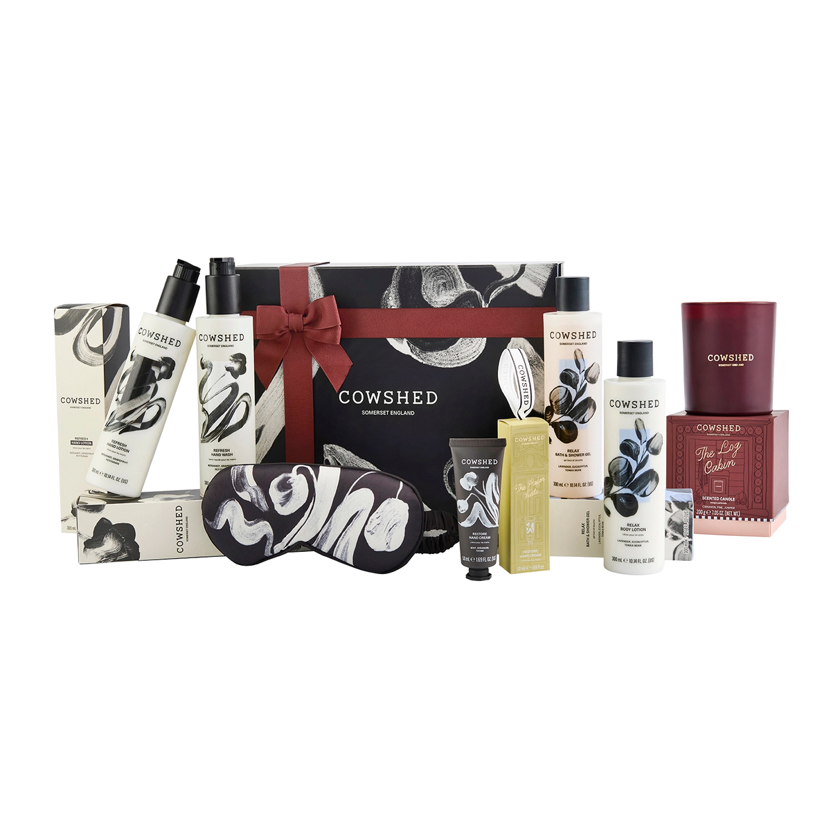Cowshed gift set featuring a black and white patterned magnetic gift box with a red ribbon, surrounded by matching bath and body products including bottles, a sleep mask, a candle, hand care all arranged on a white background.