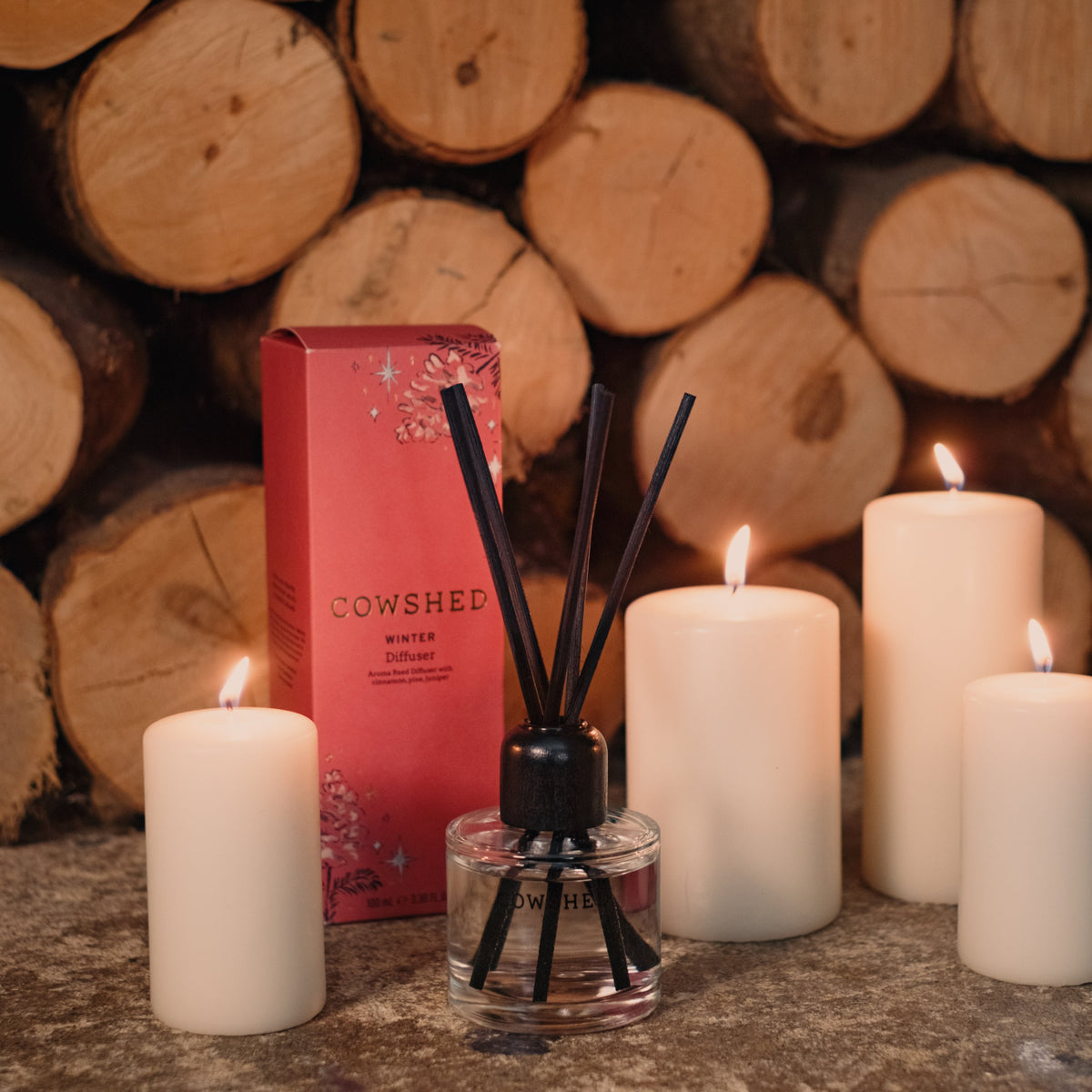 Cowshed diffuser with lit candles and wooden logs in the background
