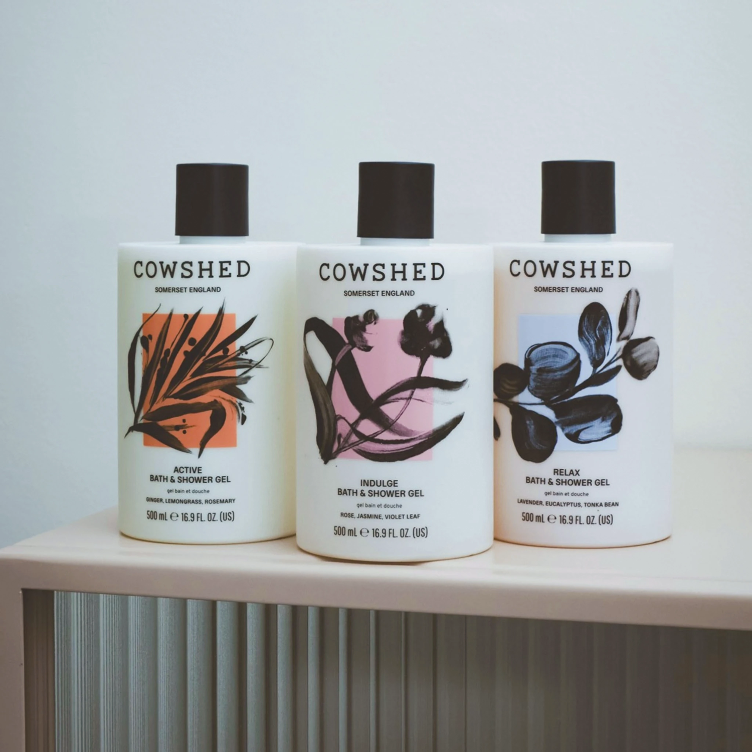 Three white bottles of Cowshed Summer of England bath & shower gel arranged on a light wooden shelf against a pale wall. The bottles have black caps and are labeled Active, Indulge, and Relax, with orange, pink, and blue botanical illustrations on the front.