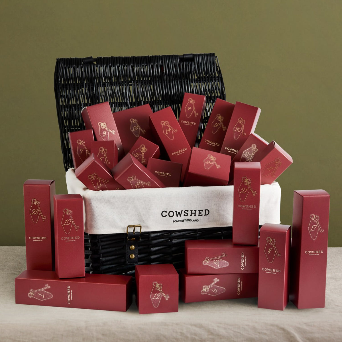 A black hamper gift basket with a white front panel printed with the brand name 'COWSHED' sits filled with and surrounded by matching dark red product boxes with gold logo graphics. The arrangement is photographed on a light fabric surface against an olive-green background.