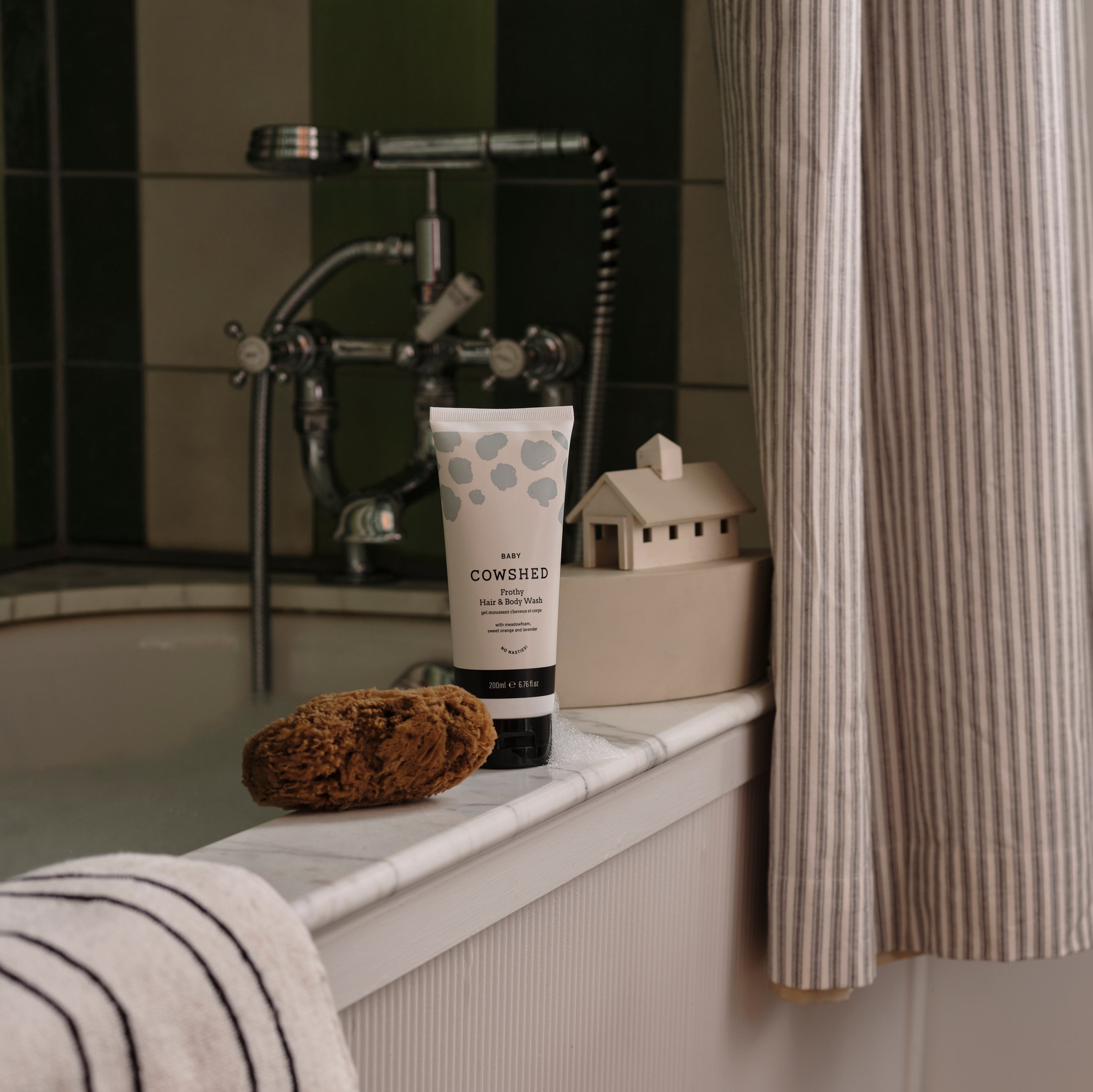 Cowshed product on a bathroom shelf with a bathtub and shower curtain in the background