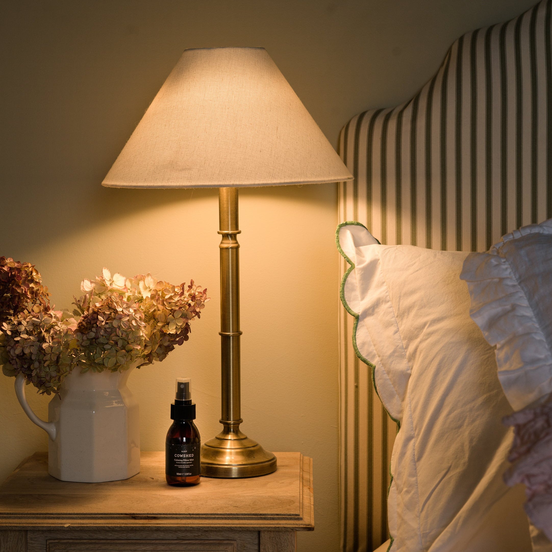 Lamp on a bedside table with flowers and a bottle, next to a bed with white bedding.