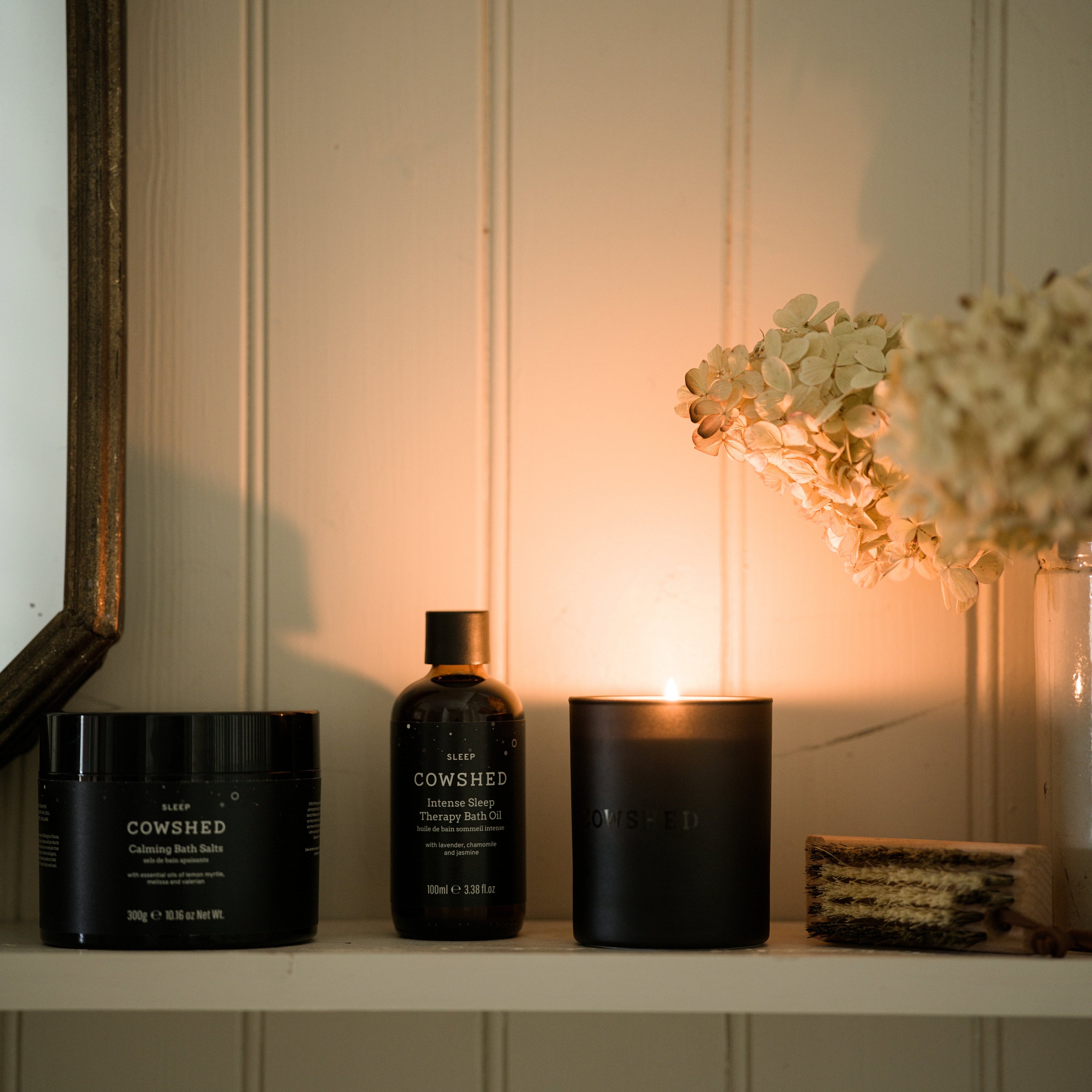 Cowshed products including a jar, bottle, and candle on a shelf with a soft light background.