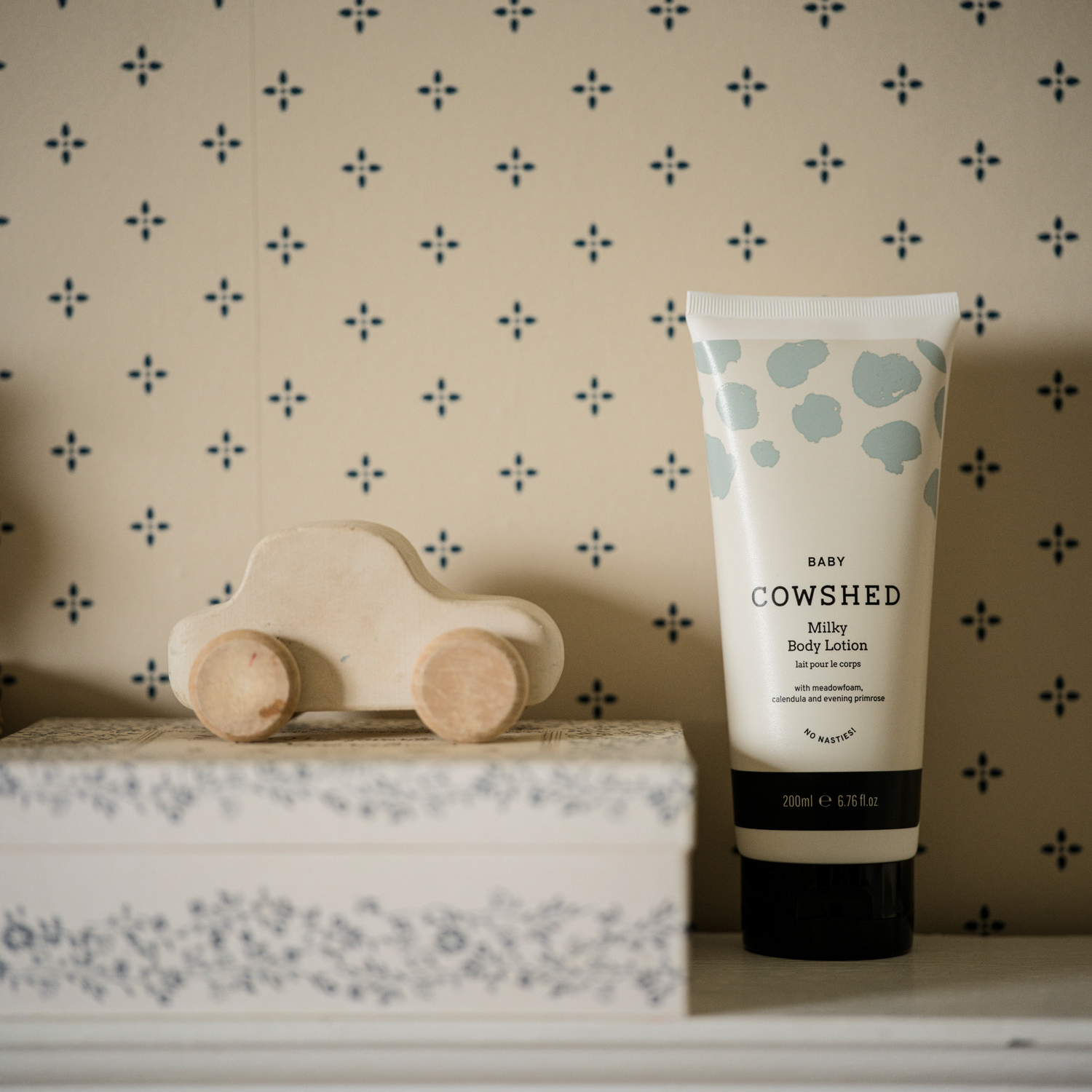 Cowshed baby body lotion with a wooden toy car on a shelf against a patterned wall.