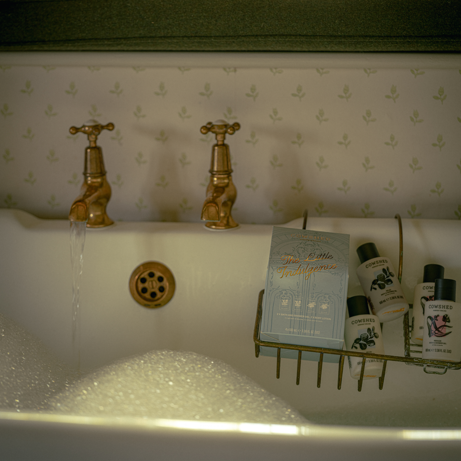 A bathroom scene with a white sink, brass faucet handles, running water, foam, and a small gold wire bath accessory rack holding cowshed toiletry bottles.