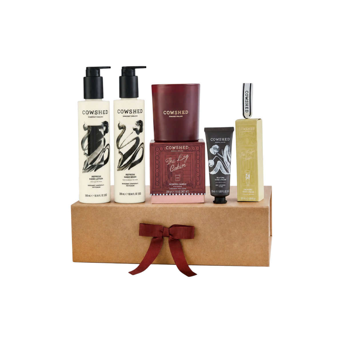 A Cowshed gift set arranged on a brown box with a dark red ribbon, featuring two hand care bottles, a candle, a hand cream tube against a plain white background.