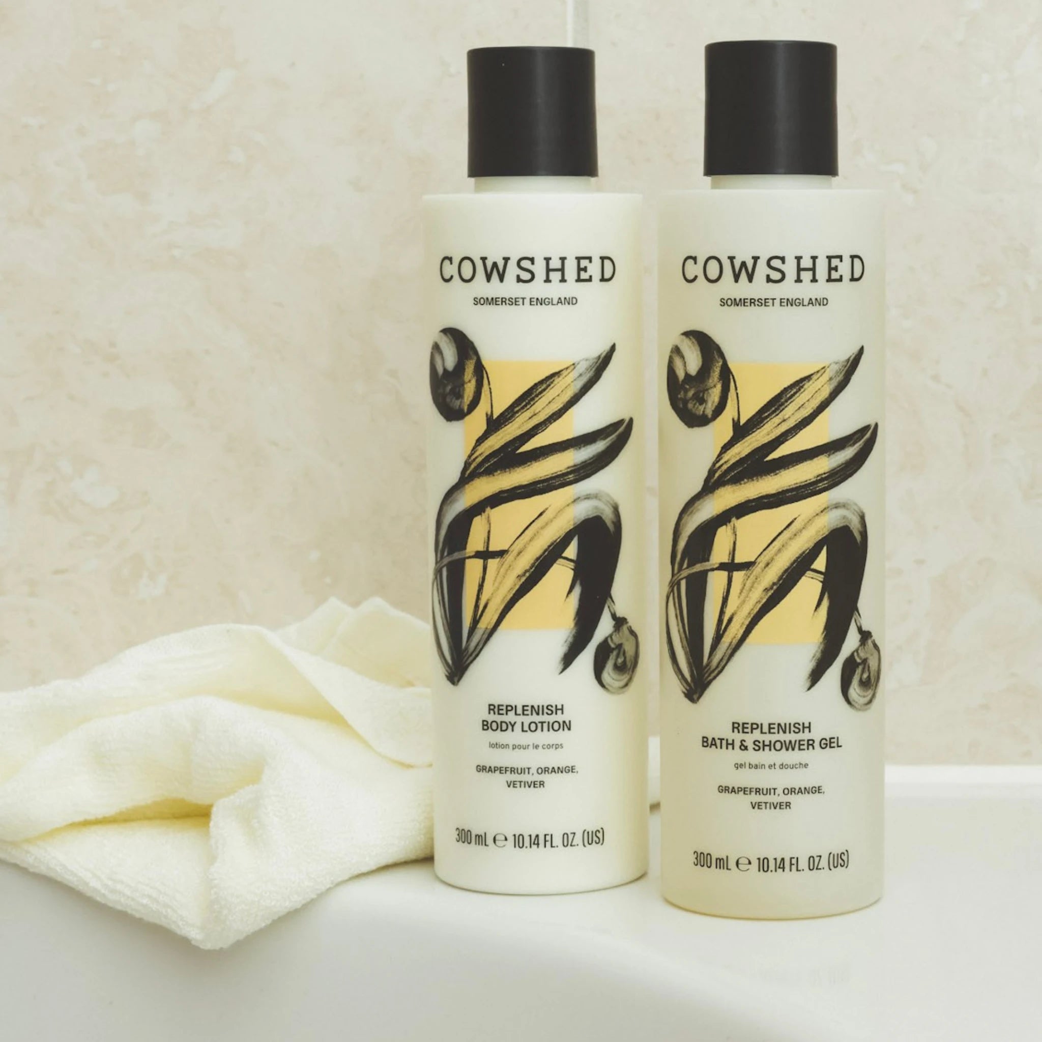Two Cowshed bottles of Replenish Body Lotion and Bath & Shower Gel on a white surface with a beige towel.