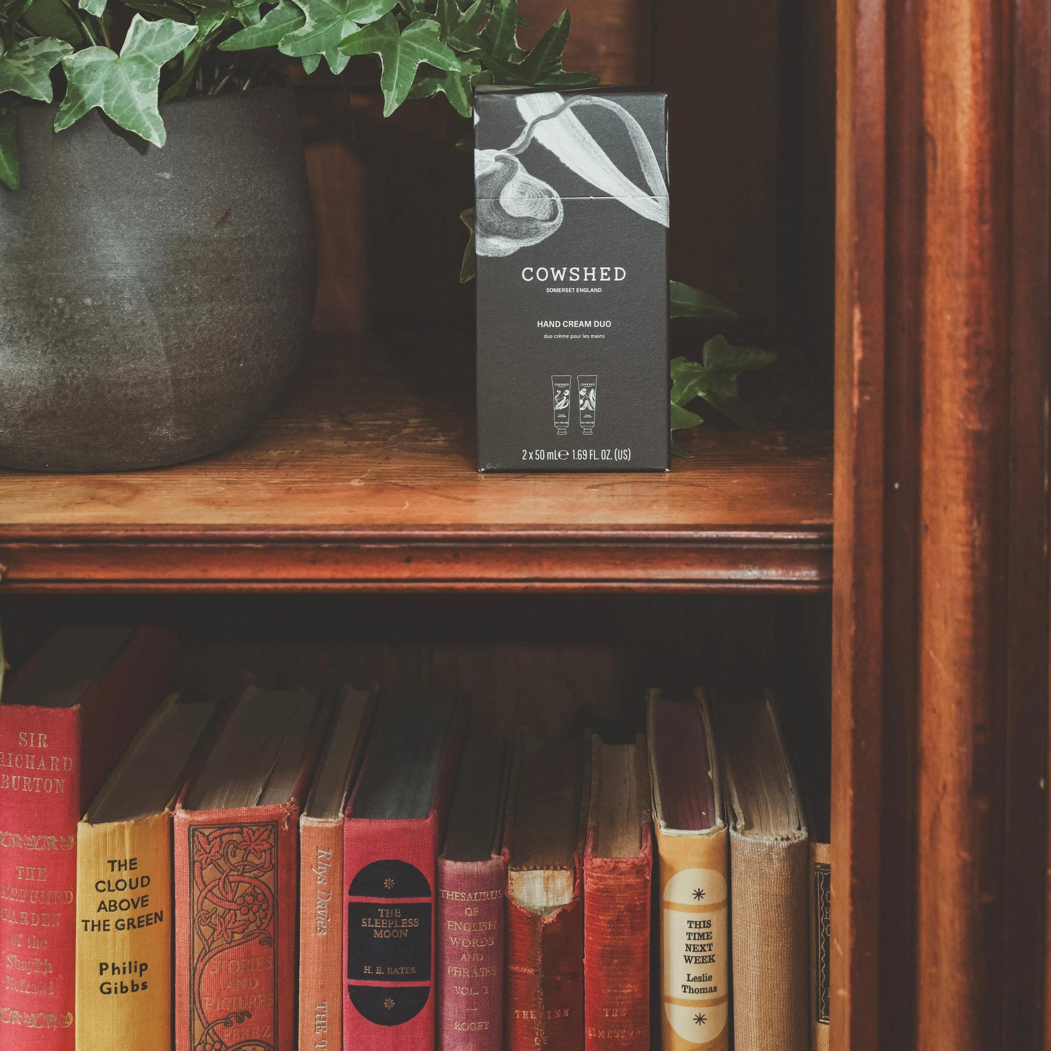 Cowshed hand cream duo on a wooden bookshelf with a plant