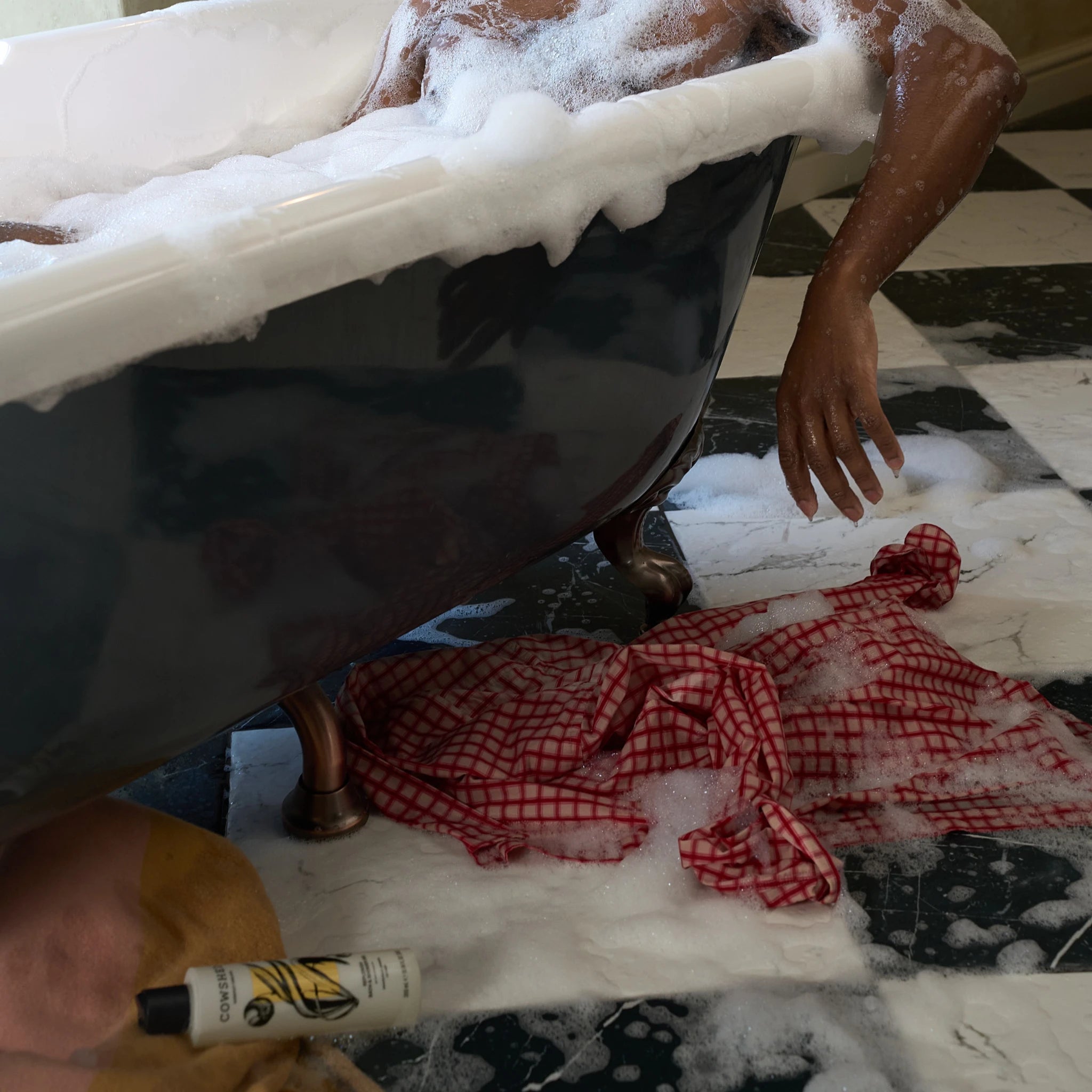 Person in a Cowshed bath with soapy hands and a red checkered towel on a checkered floor