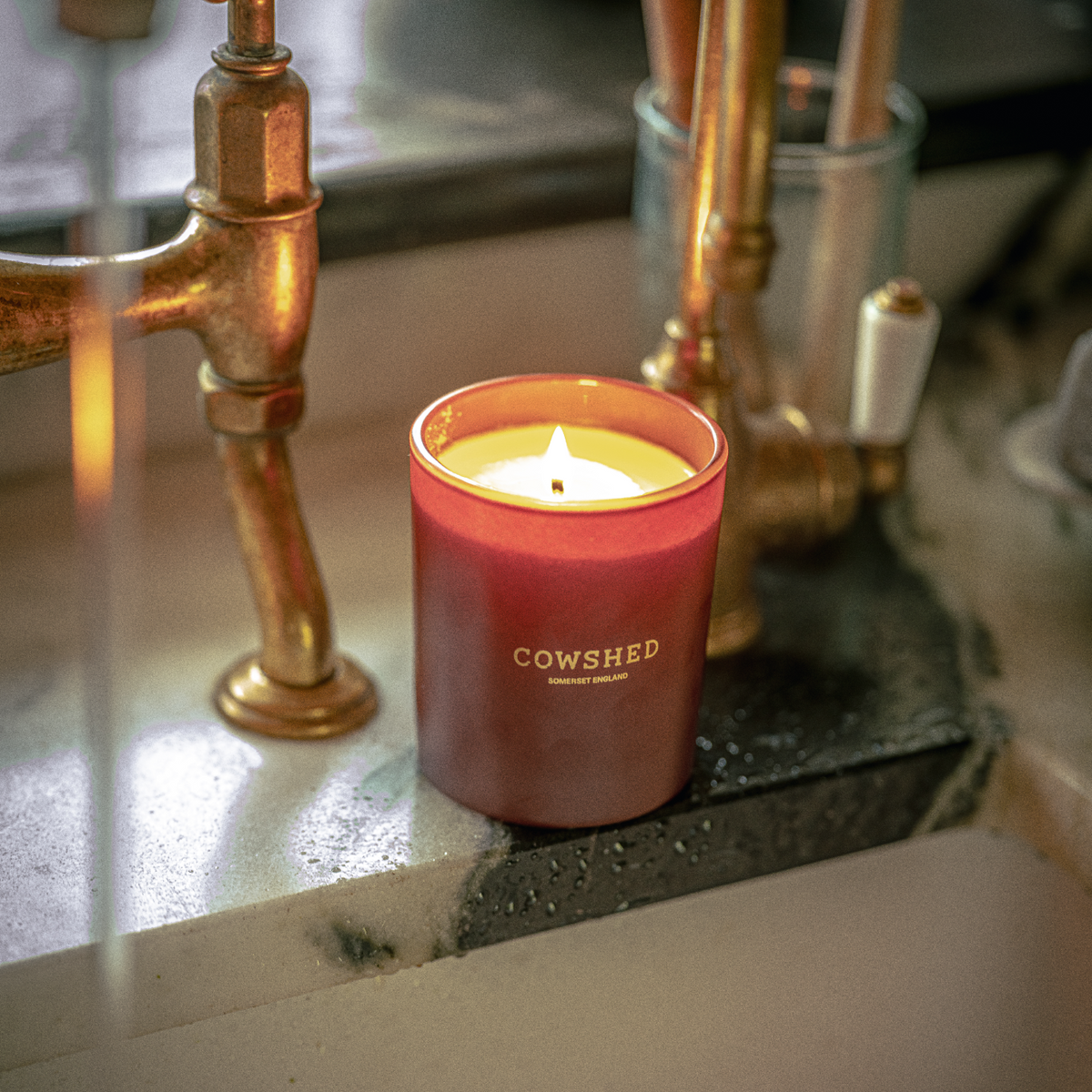 A lit winter candle in a matte terracotta red jar with the brand name 'COWSHED' printed in gold on the front, set on a kitchen sink next to brass fixtures.