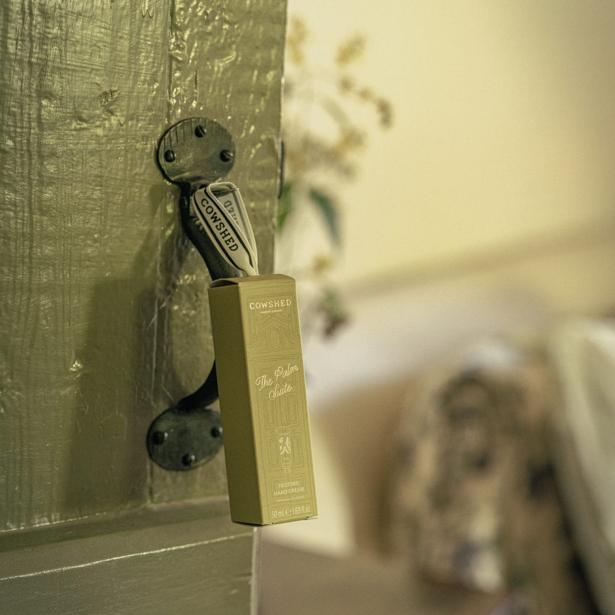 A Cowshed hand cream bauble Christmas gift hangs from a black metal latch on a rustic green wooden door, with a warm, softly blurred interior background.