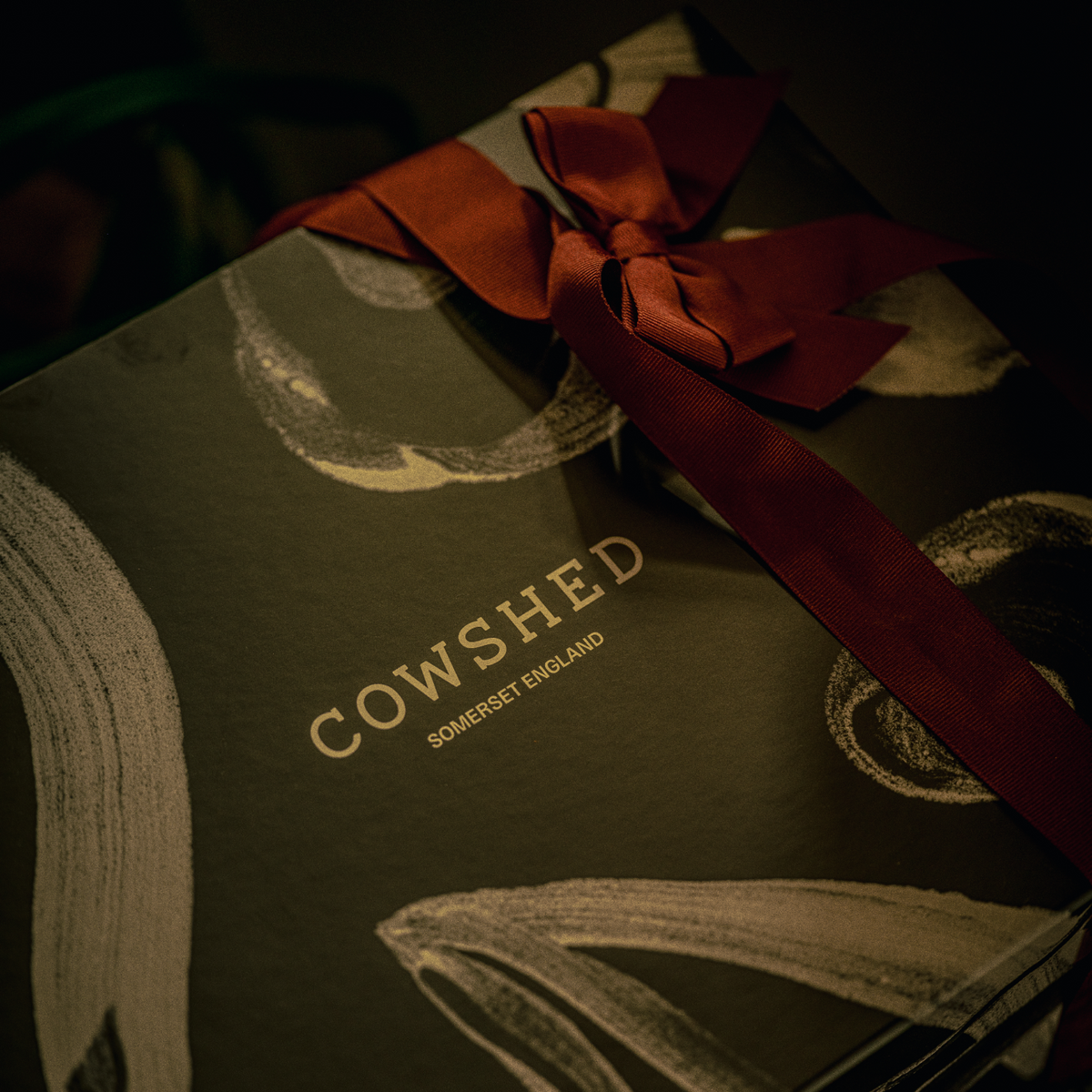 A close-up of a Cowshed gift box or package in dark olive green with gold brushstroke patterns and a large dark red ribbon tied in a bow. The brand name 'COWSHED' and 'SOMERSET ENGLAND' are printed in gold on the front.