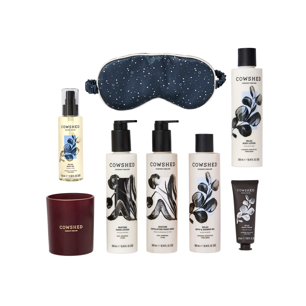 A Cowshed self-care gift set featuring a navy blue speckled sleep mask, a navy blue candle, a bottle of lavender body wash, a bottle of body lotion, two bottles of shower gel, and a black tube of hand cream, all arranged on a clean white background.