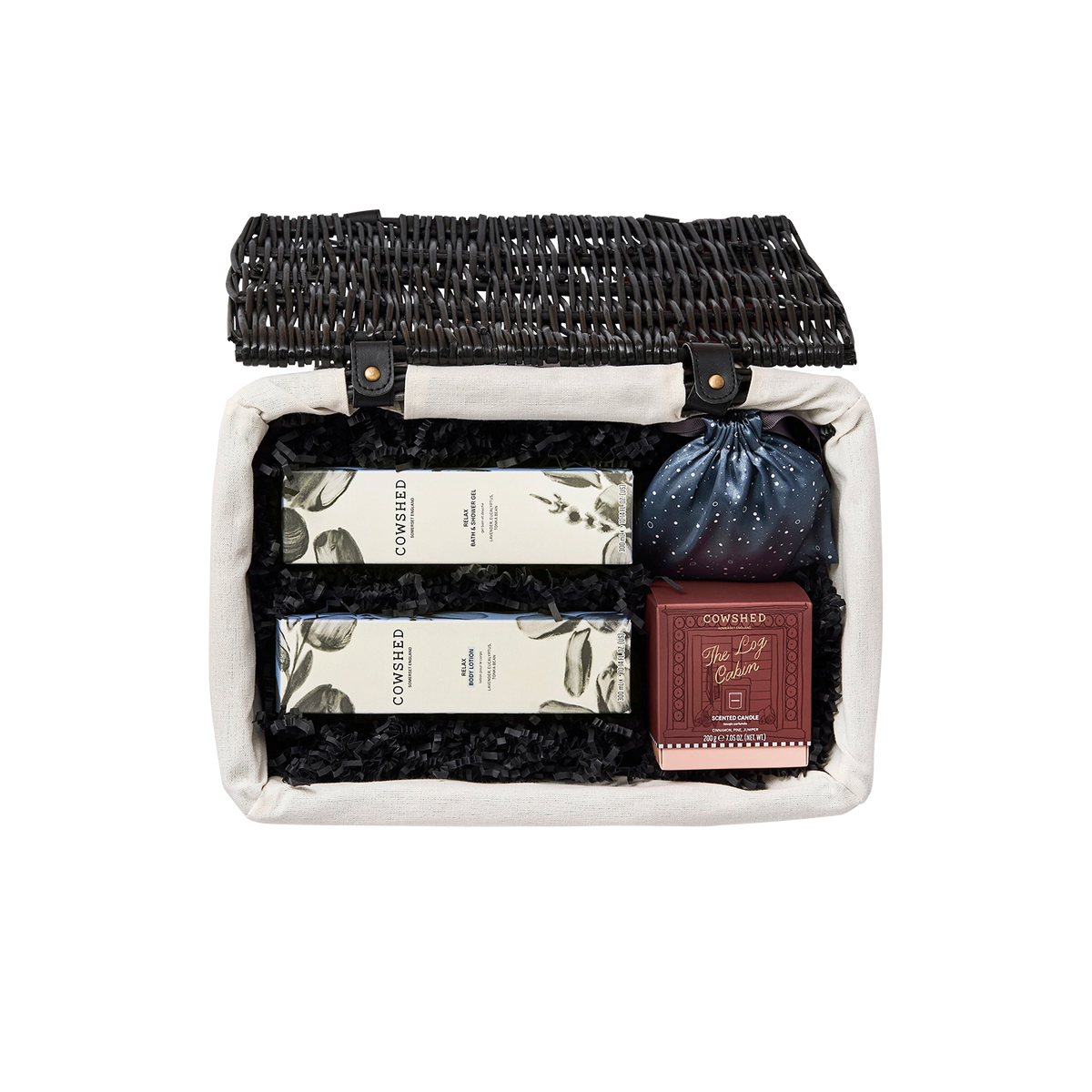 An open wicker gift basket with a cream fabric liner and black crinkle paper filler, containing two boxed items with botanical leaf prints, a small dark blue drawstring pouch, and a reddish-brown boxed item. The brand name 'COMBINED' is visible on the products.