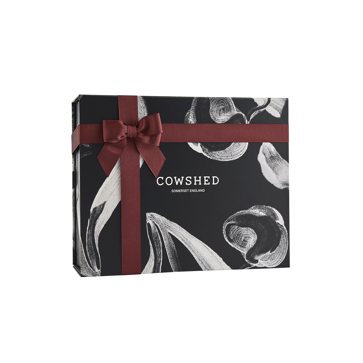 A rectangular Cowshed gift box in black with white abstract shell illustrations and a dark red ribbon tied in a bow across the top, shown on a plain white background.