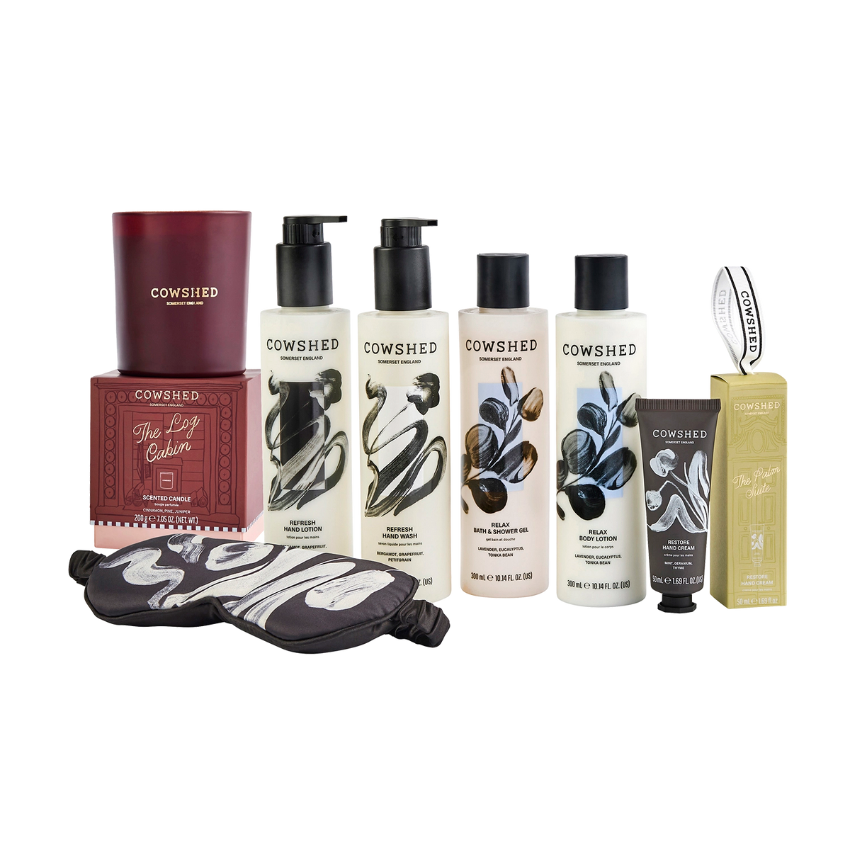 Cowshed beauty gift set featuring a maroon candle in a box, four white hand and body care bottles with black caps, a black  cowshed eye mask, and a gold-colored box, all arranged on a white background.