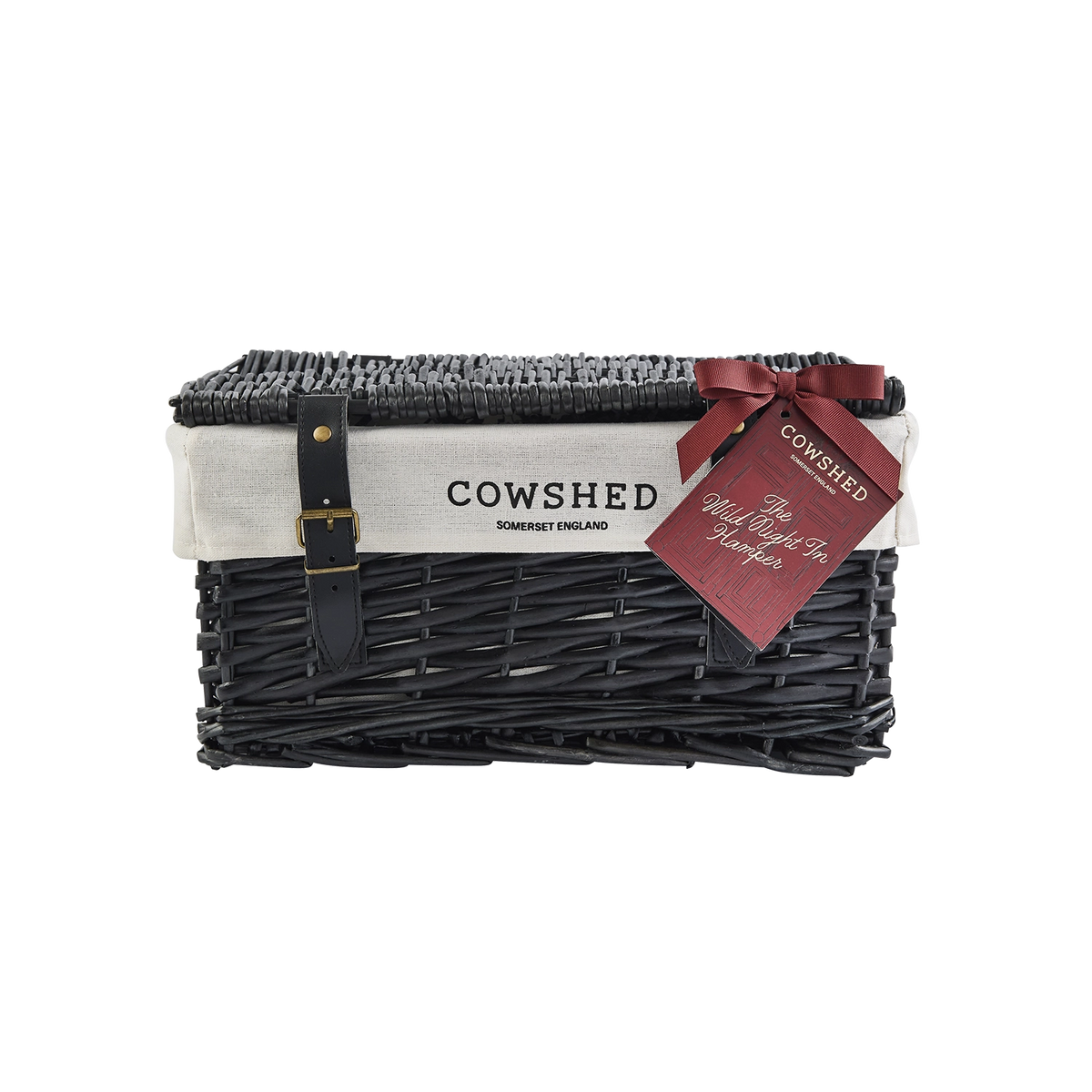 A black wicker gift basket with a white paper band reading 'COWSHED Somerset England' and a dark red ribbon bow with a matching tag, photographed on a plain white background.