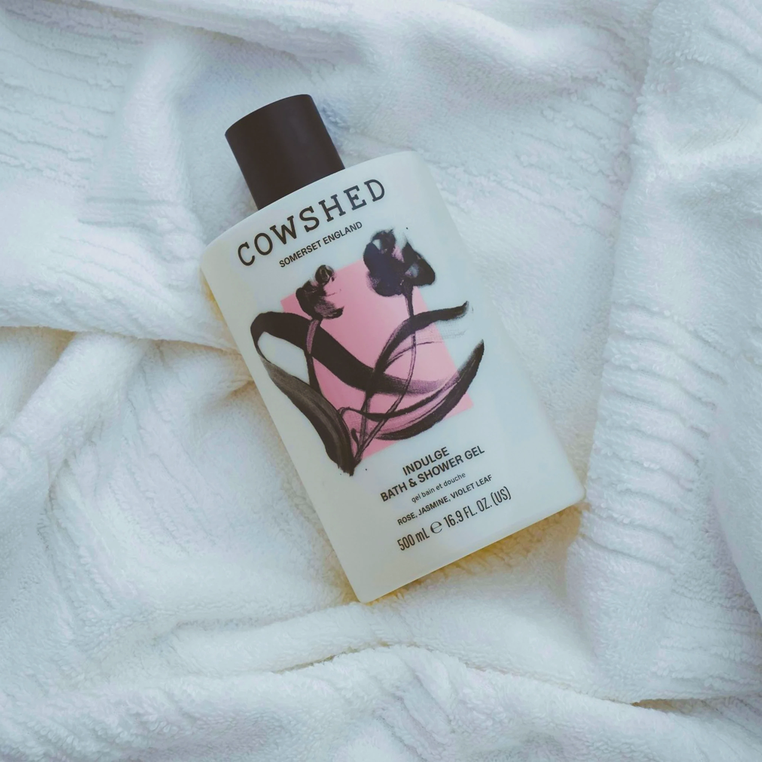 A bottle of Cowshed Indulge Bath & Shower Gel placed on a soft white textured towel. The bottle is frosted with a black cap and features a label with a pink background and black botanical illustrations.