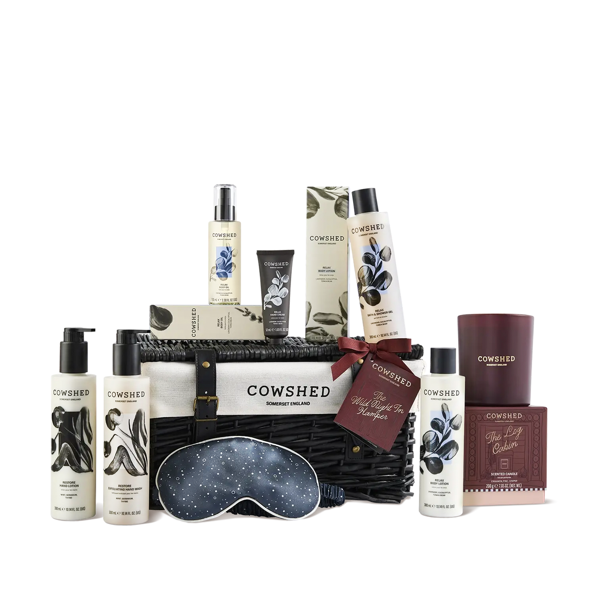 A Cowshed gift basket set featuring multiple bath and body products, a candle, a sleep mask, and a box, arranged in a black woven basket on a white background.