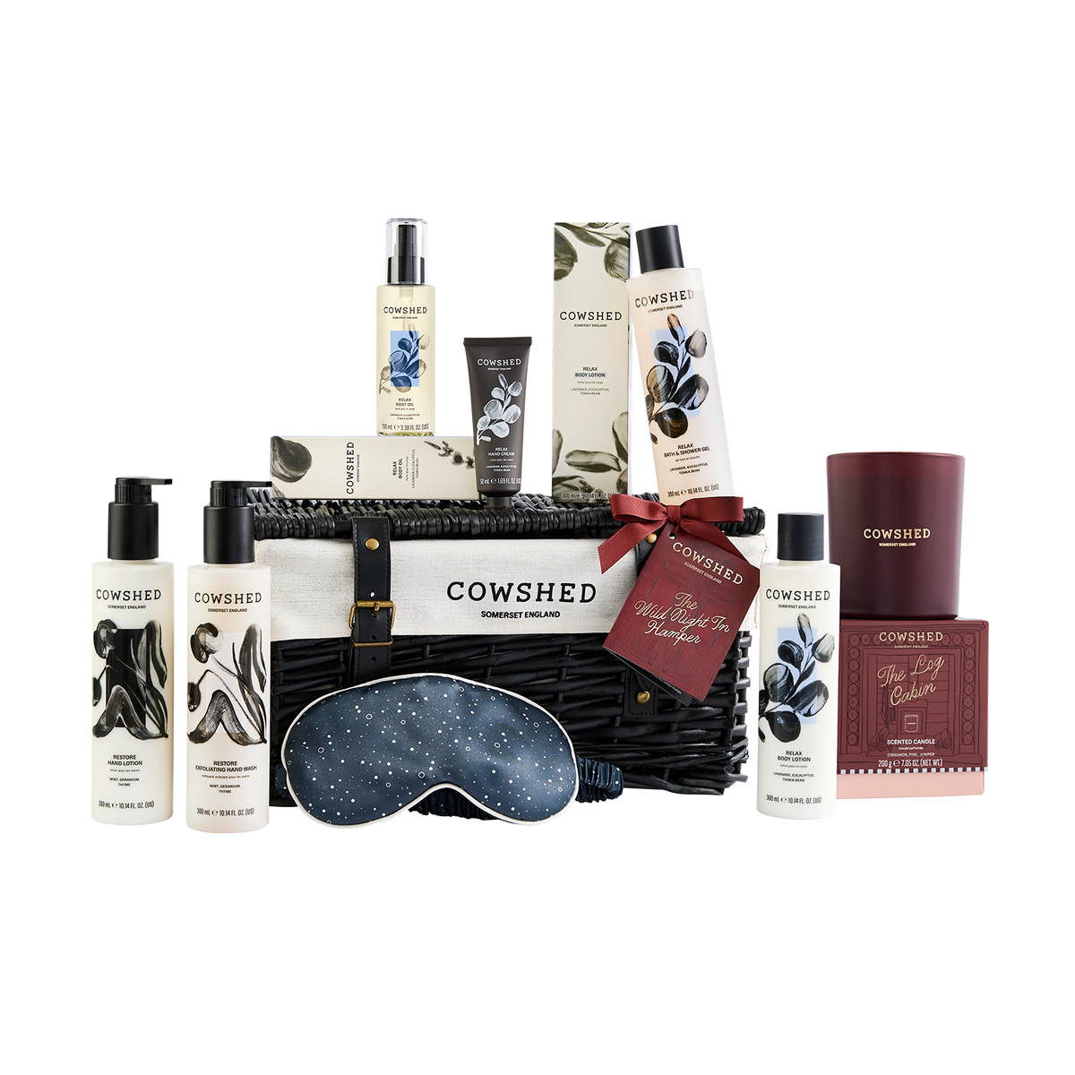 Cowshed Self Care Gift Hamper featuring a black wicker basket filled with bath and body products, a navy blue starry sleep mask, and a dark brown candle, all on a white background.
